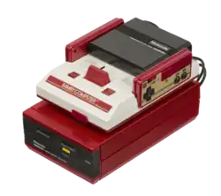 A white and red Famicom unit sits atop a candy red Famicom Disk System unit with black insertable disk drive. Two rectangular controllers, each with a D-pad and two black buttons, fit into the Famicom.